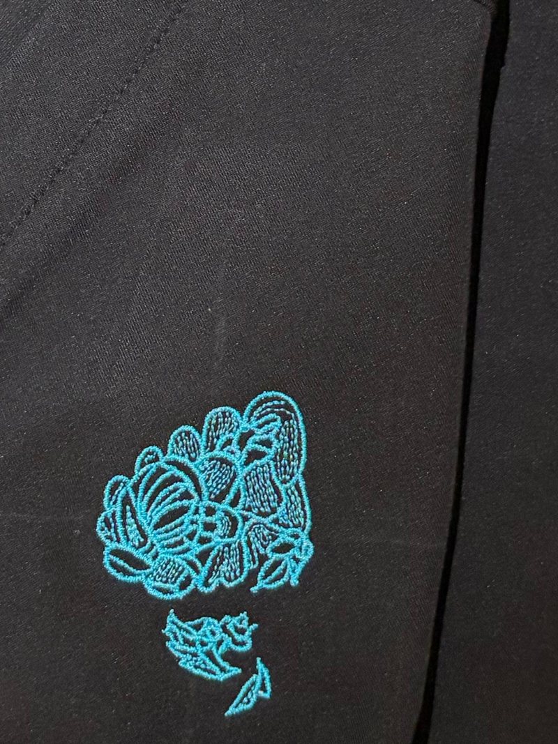 Close-up of the LoveFeet logo embroidered in turquoise thread on a black Stitchwell scrub top